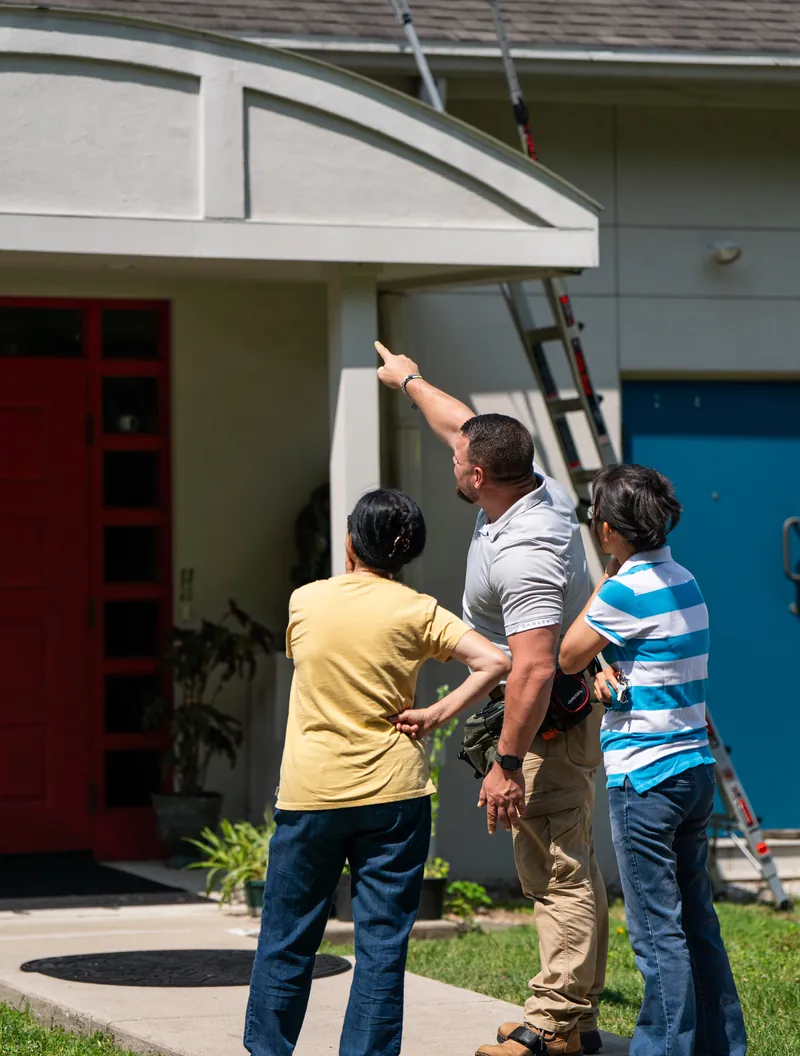 A roofer pointing up at a roofline while two homeowners look up at it with him.