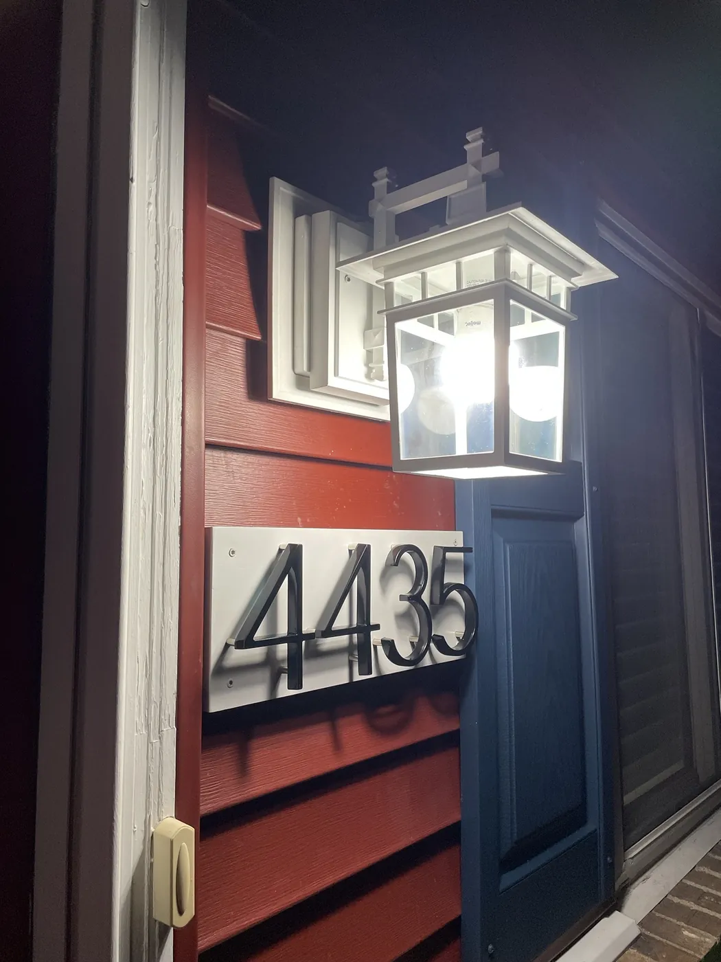 Red vinyl siding with white trim and a blue shutter beside a lit porch lantern.