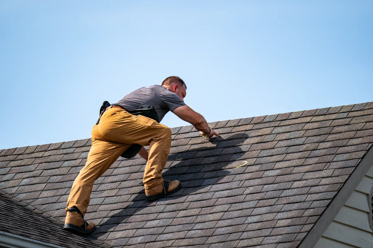 Travis Haessly working up the slope of a shingle roof, reaching toward a chalk mark on the shingles.
